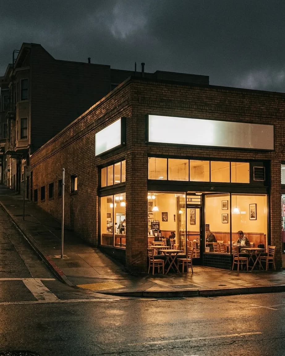 A caffè front on a corner at night with warm window light on damp pavement and a steep street rising to one side