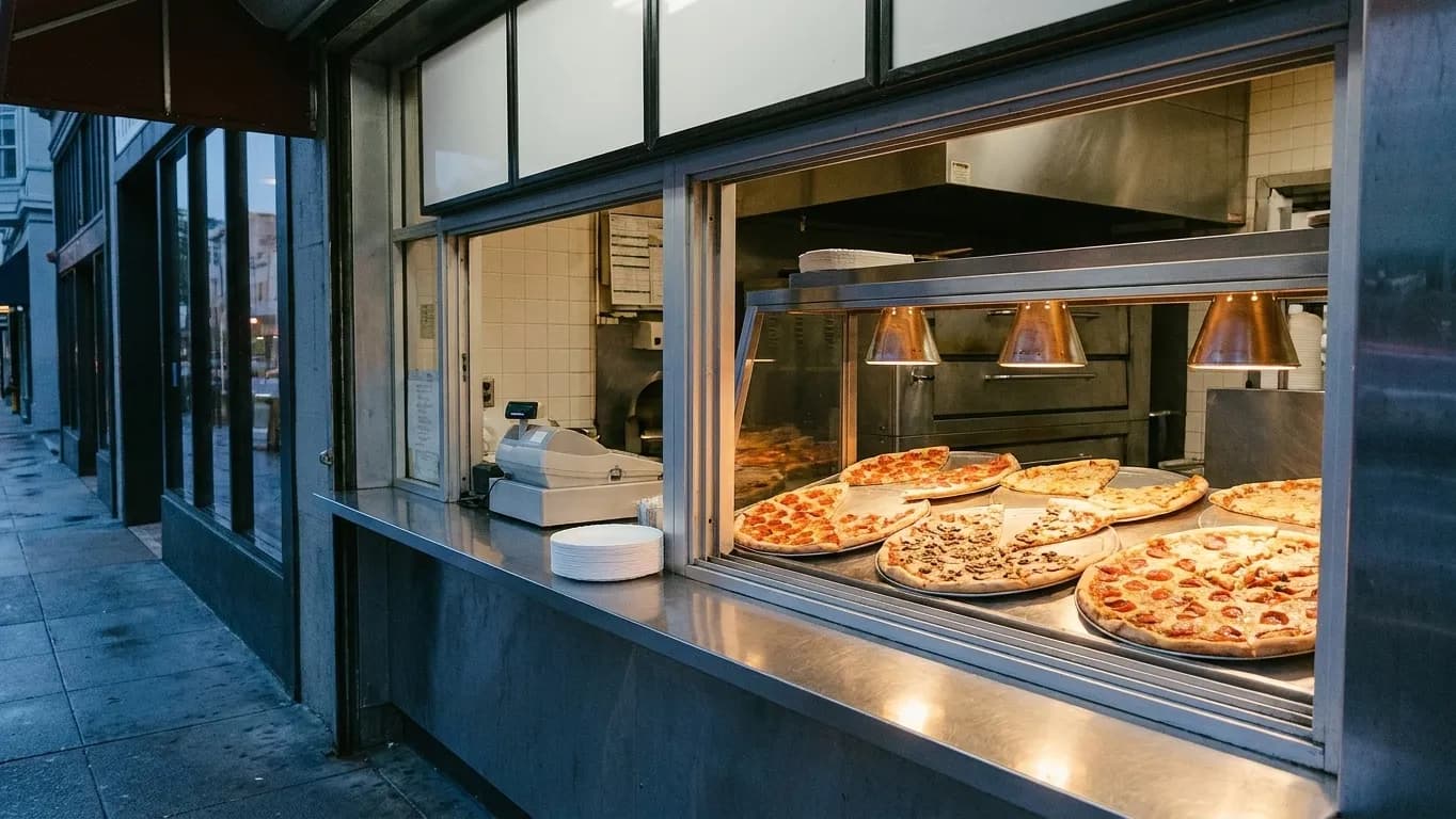A San Francisco slice counter seen from the sidewalk at dusk, with warming trays under a heat lamp and a stack of paper plates