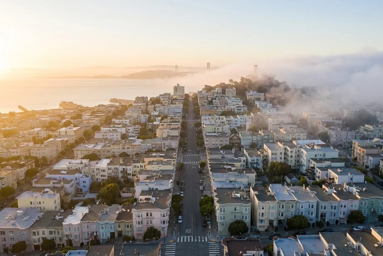 A San Francisco hilltop view of pastel houses stepping down a steep grid, fog spilling over the ridge on one side while the other stays in gold light