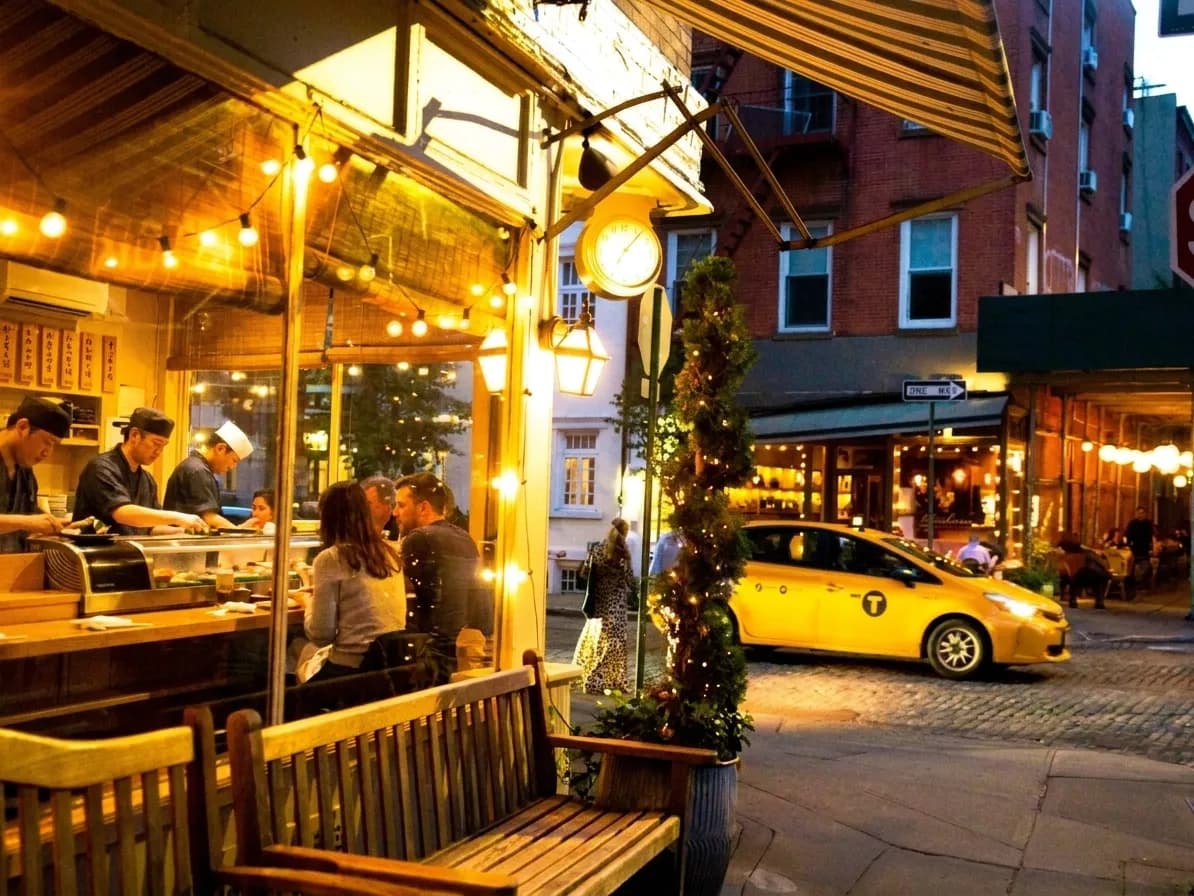 Night exterior of a small sushi counter glowing on a cobblestone Greenwich Village corner as a yellow cab passes
