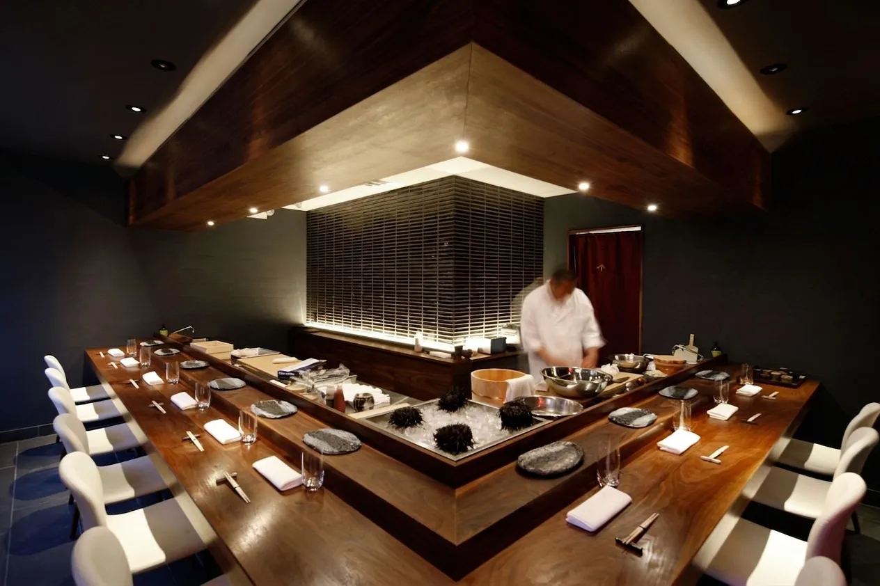 Warmly lit wooden omakase counter interior with a chef in whites working behind trays of ice and sea urchin