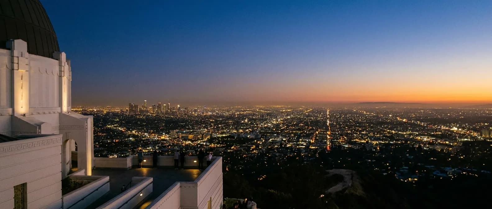 A hilltop observatory building at dusk with the city lights spreading out below and a few silhouetted visitors on the terrace
