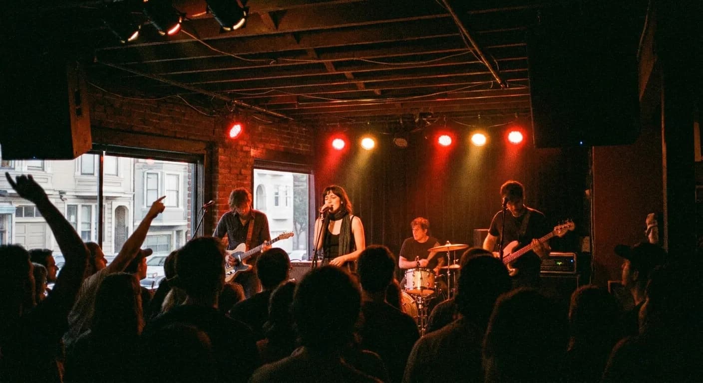 A small dark music venue from within a modest crowd, a band lit in red and amber on a low stage close by, silhouetted heads and raised arms in the foreground