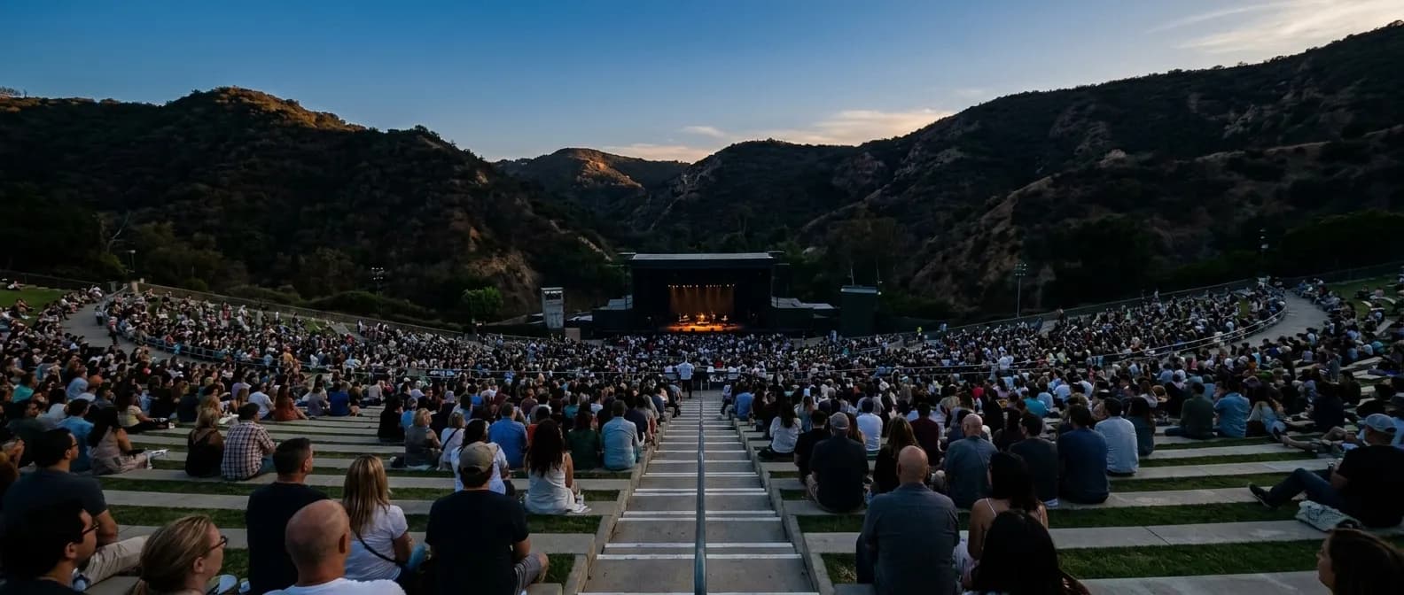 A large outdoor amphitheatre at dusk seen from the back rows, the stage lit far below and the crowd filling in