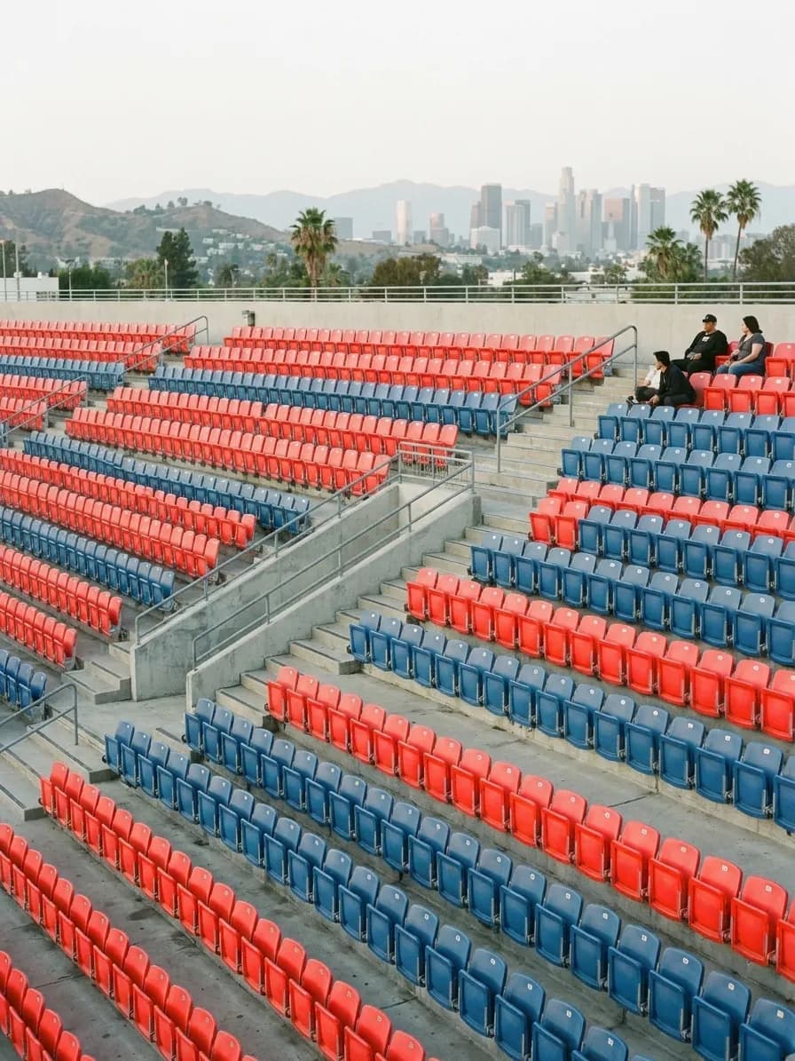 Rows of empty coloured plastic stadium seats photographed straight on in flat afternoon light, a repeating geometric pattern with a few people seated far apart
