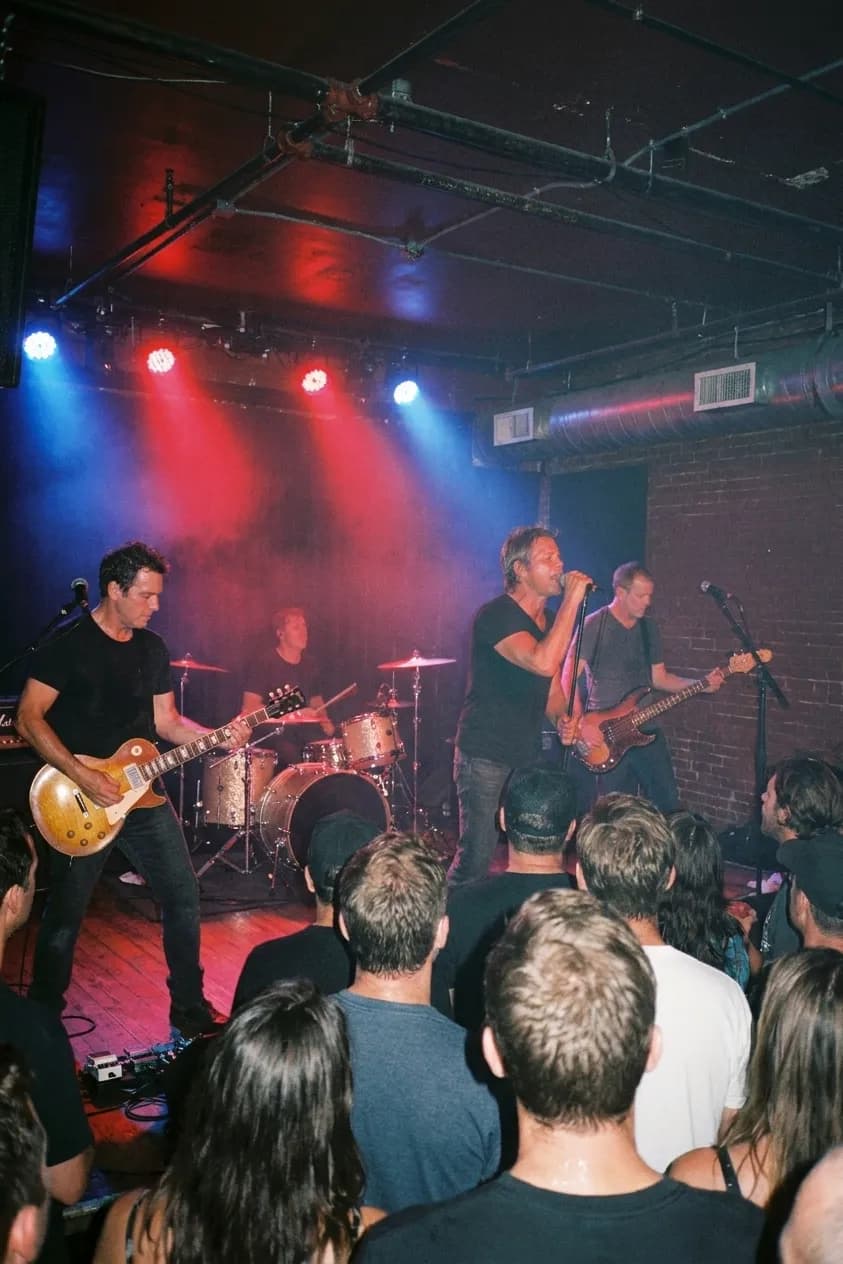 A band performing on a small club stage seen from the middle of a standing crowd