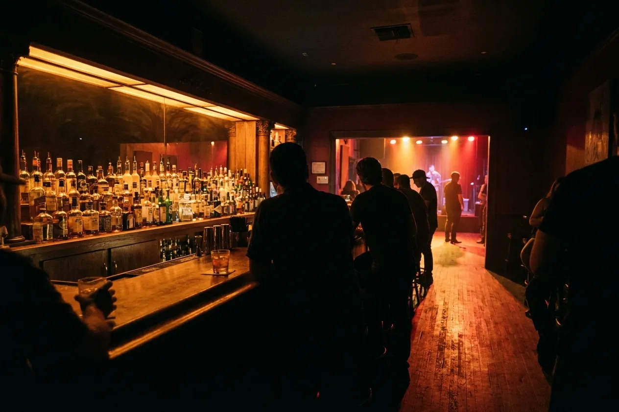 The interior of a dimly lit bar adjoining a music room, a long counter with bottles backlit in warm amber and a few people standing with drinks in silhouette