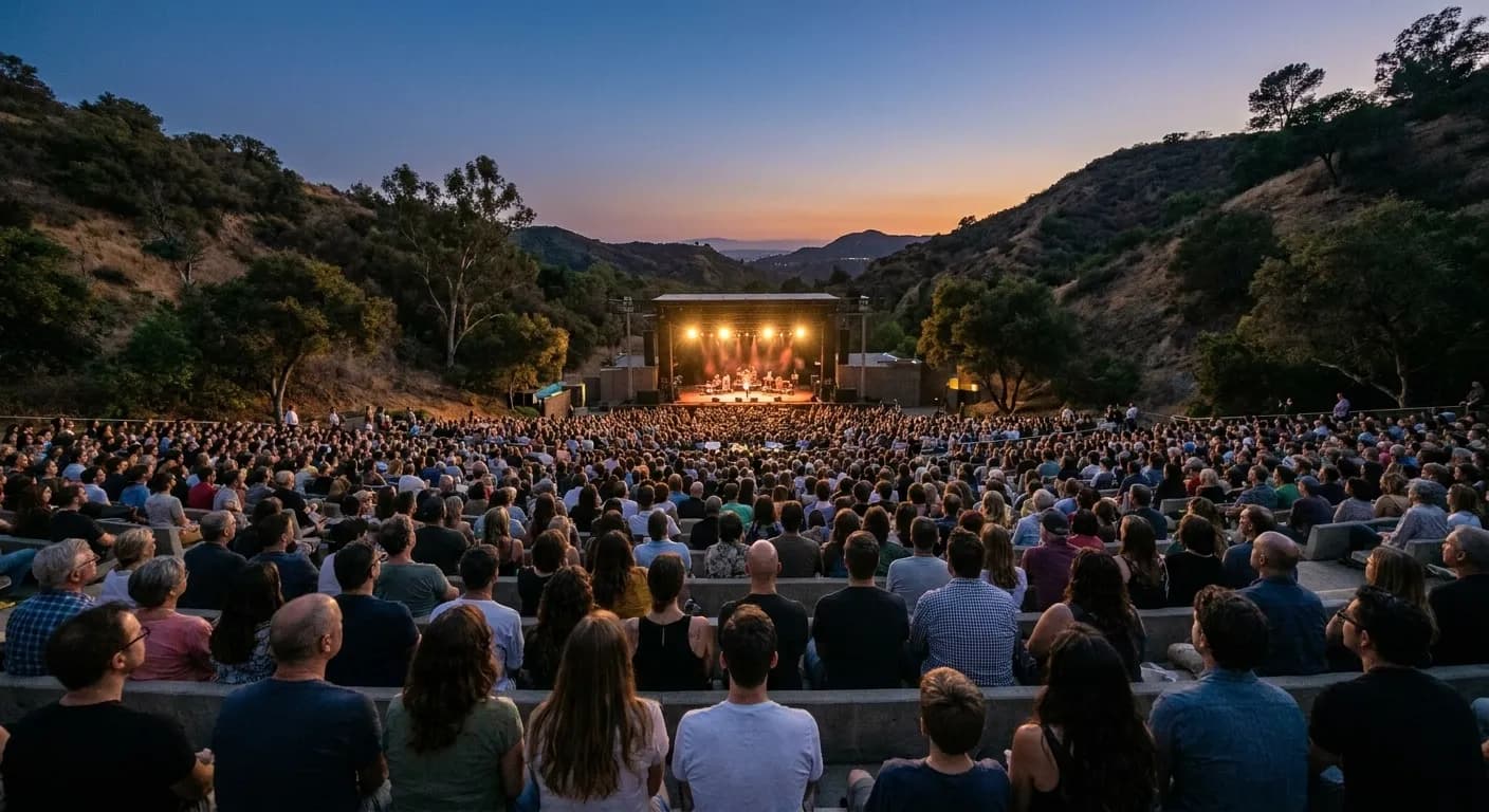 An outdoor amphitheatre audience seated on tiered benches at dusk with a lit stage below