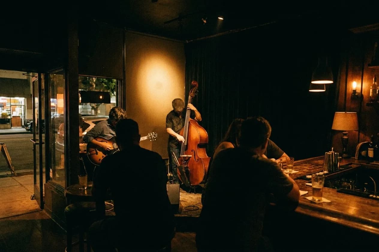 A small band playing in the corner of a dimly lit bar with drinkers nearby