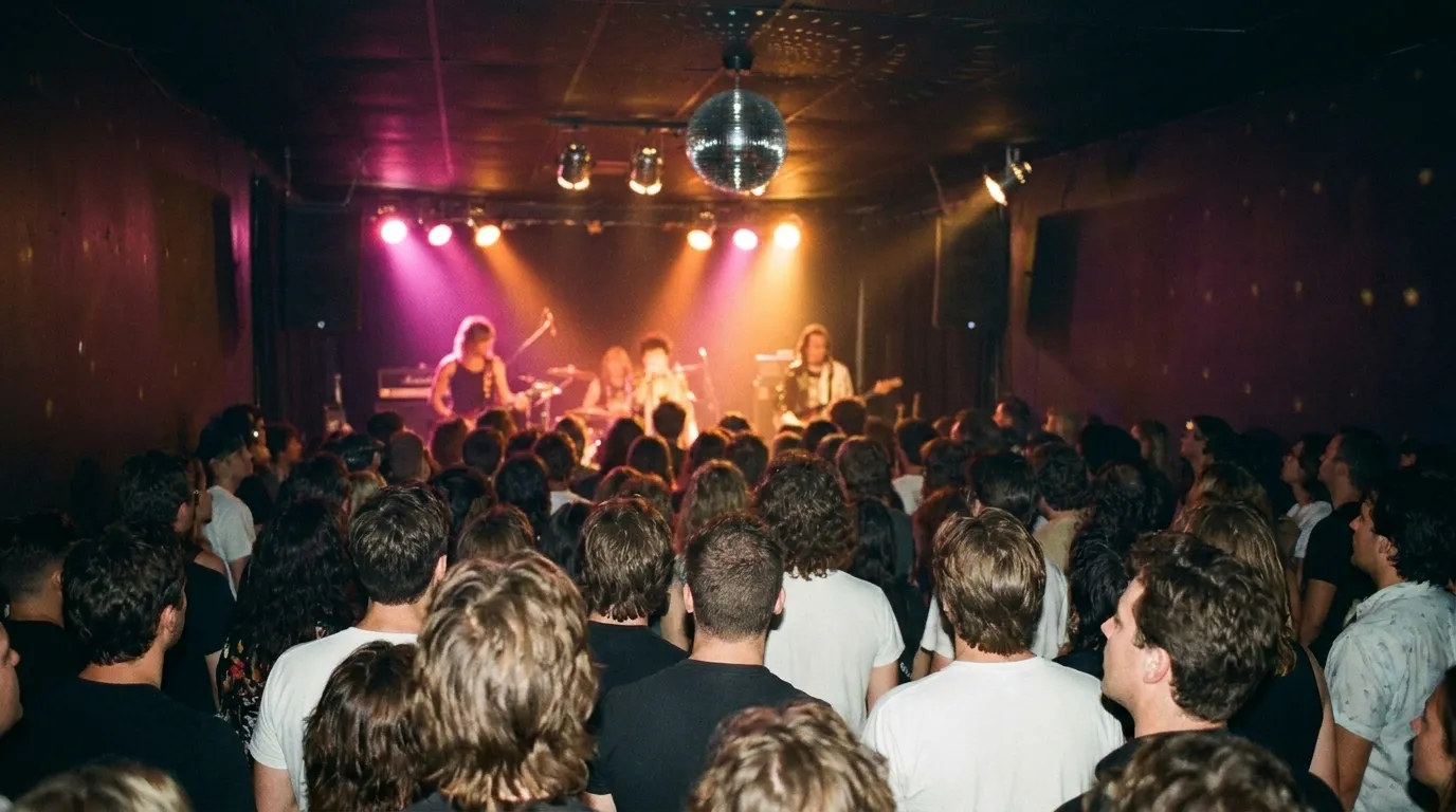 A crowd packed against a low stage in an upstairs music hall, hands up, coloured stage wash overhead.