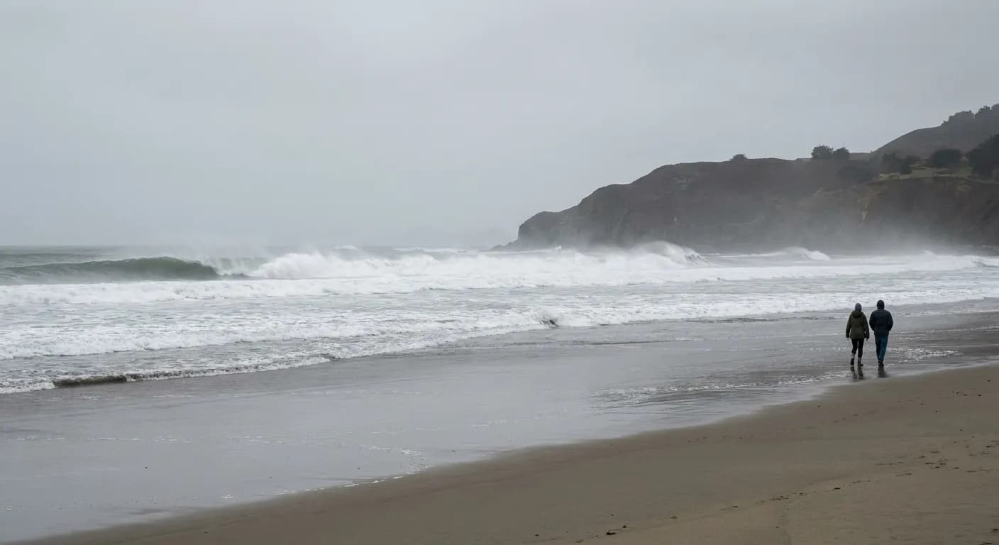 A wide empty sand beach with heavy surf and a headland in the distance under grey sky