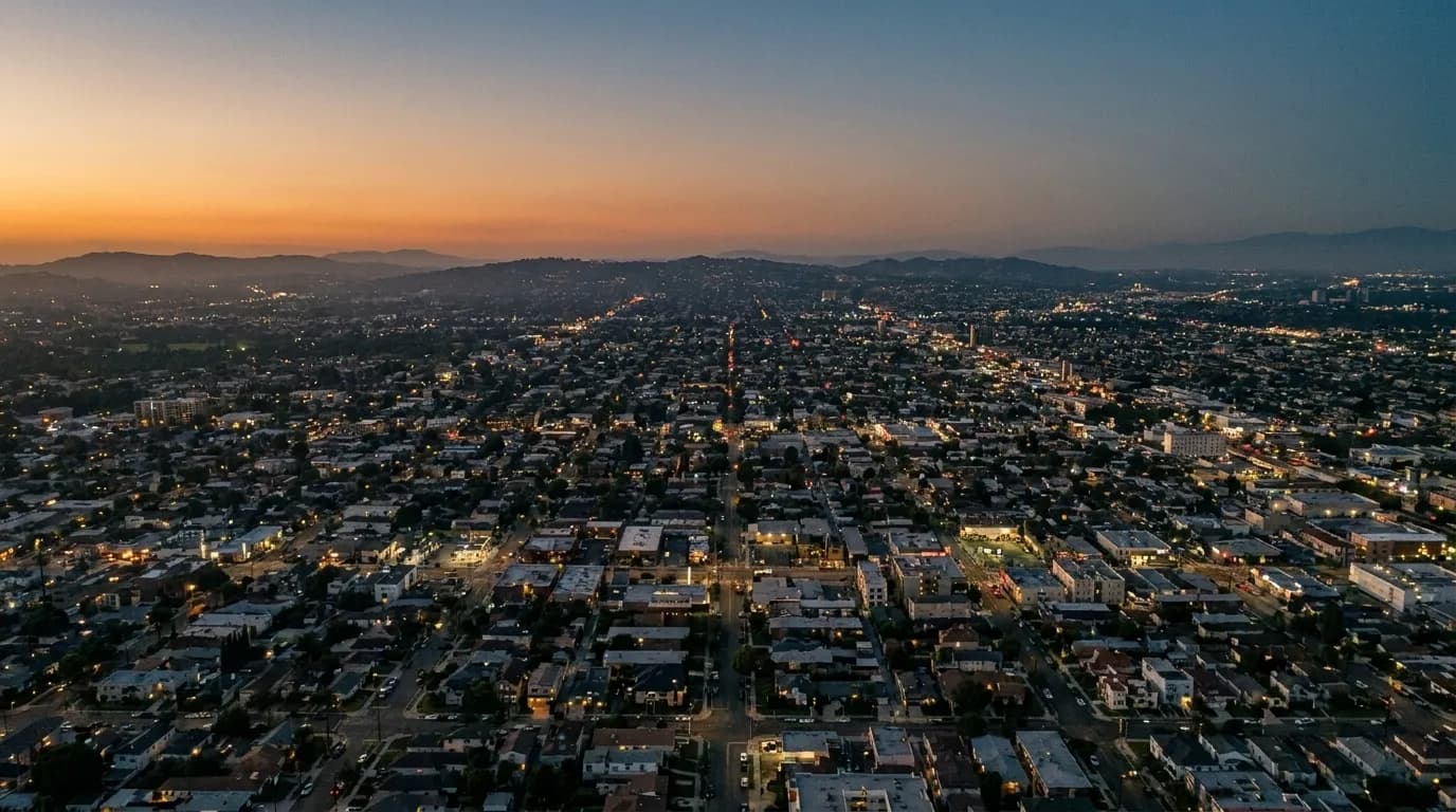 A wide city view at dusk with hills behind and lights coming on