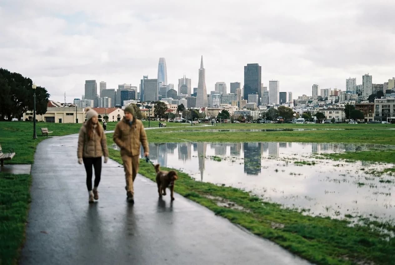 Two people walking a path through a park with the city skyline behind