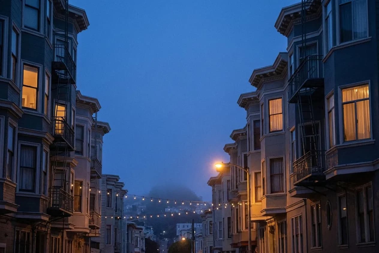 Bay-windowed San Francisco buildings with iron fire escapes framing a fog-softened hill at blue hour, cafe bulbs strung across the street
