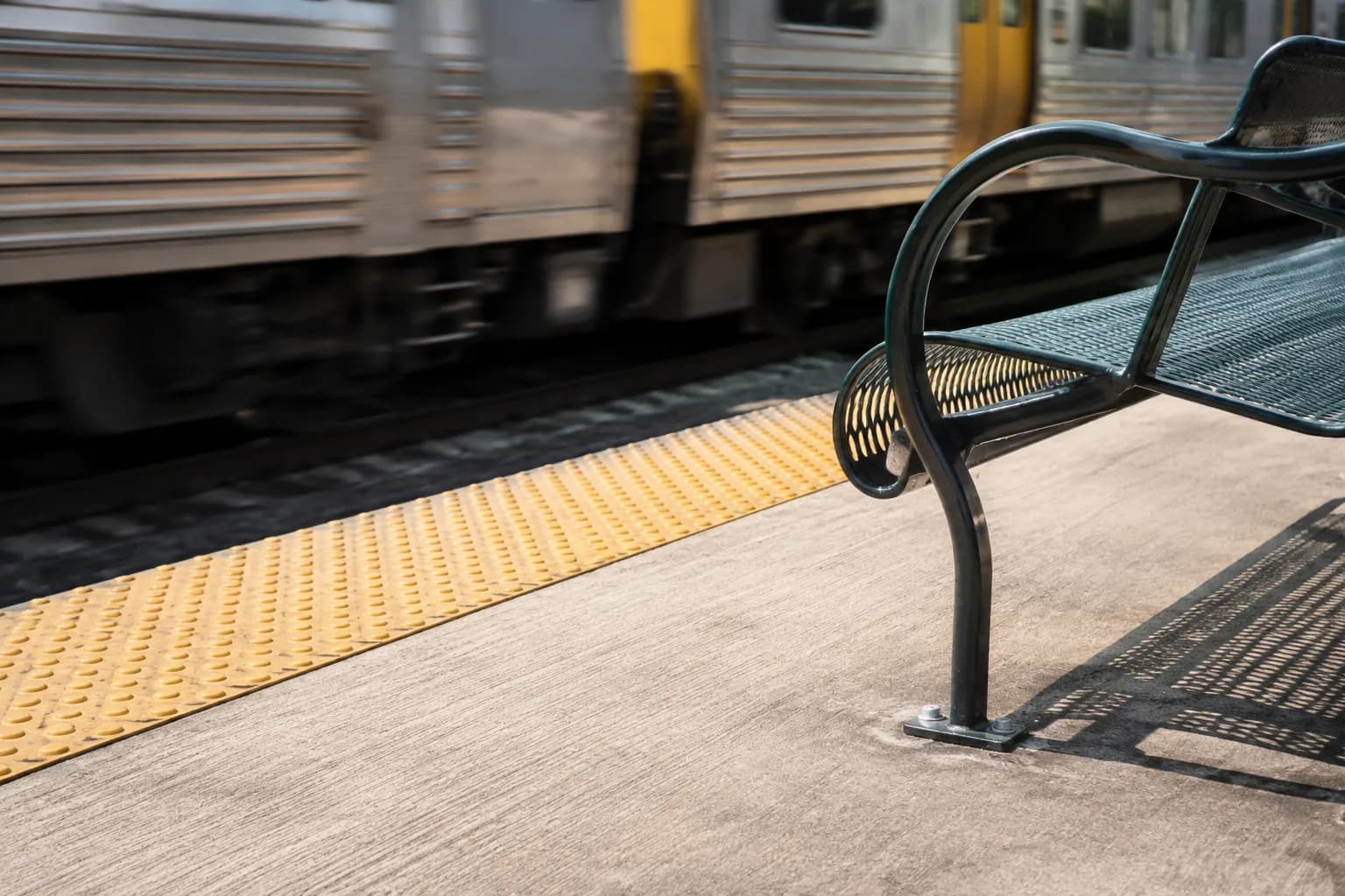 Highland Park Metro Gold Line station platform showing tactile warning strip, concrete surface, and metal bench with a train passing in the background