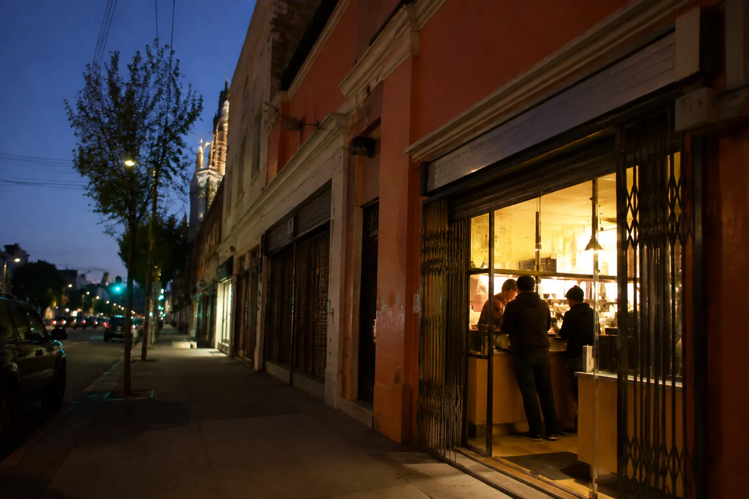 Evening view of taquería-lined 24th Street in San Francisco's Mission District with warm interior light and sidewalk foot traffic