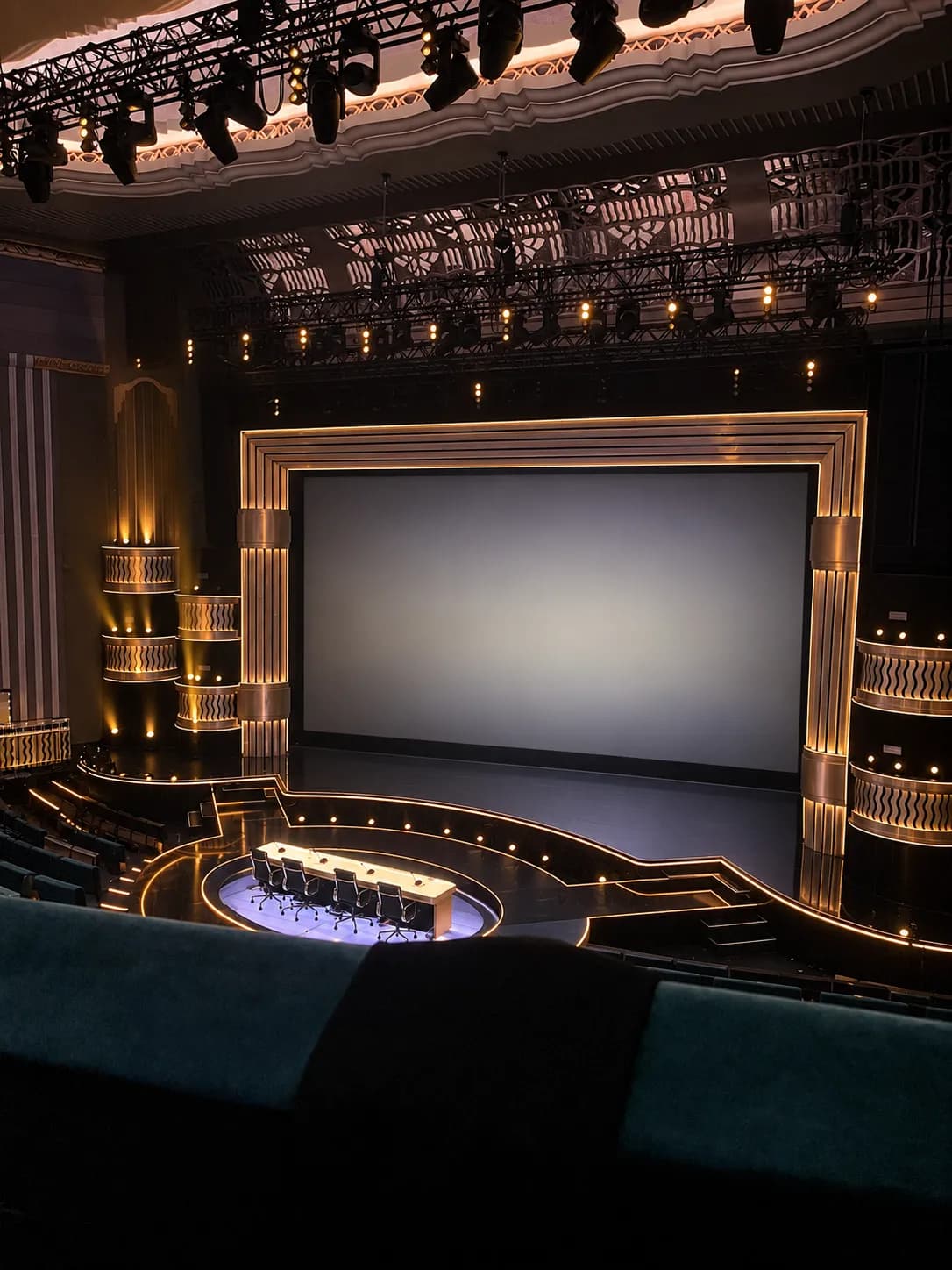 Empty cinema auditorium seen from the back row, with raked dark seating, a narrow service table along each row and a blank illuminated screen