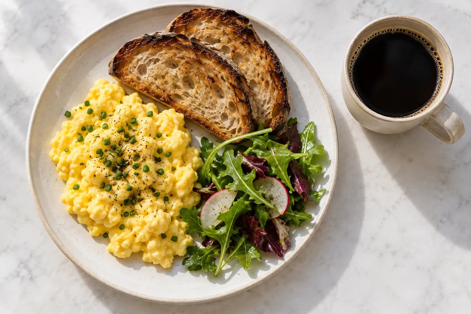 Soft scrambled eggs with chives beside thick toasted sourdough and a small salad on a pale ceramic plate, with black filter coffee alongside