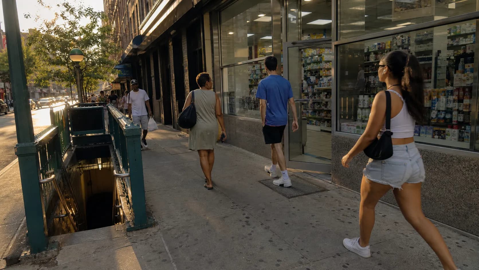 Pedestrians walk along a New York City sidewalk near a pharmacy and subway entrance.