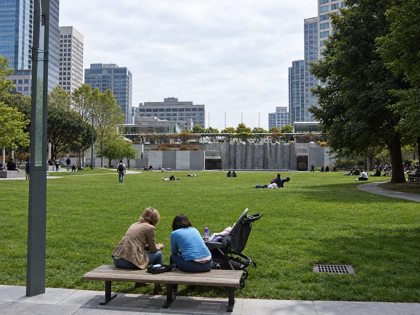 Urban public garden between tall buildings with a broad lawn, a curved water feature wall and paved terraces with benches