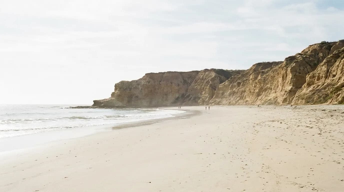 A wide pale sand Southern California beach with low surf below sandstone bluffs in hazy bright coastal light