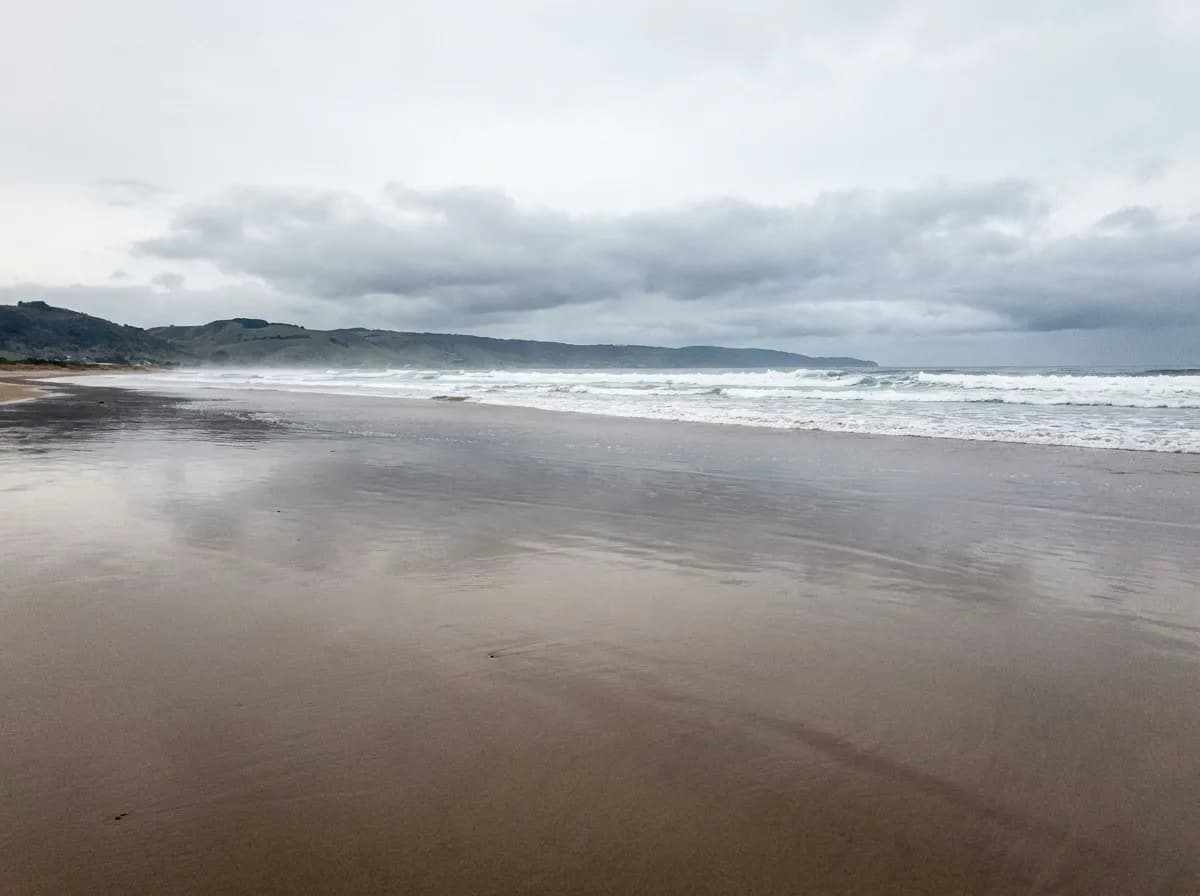 A wide empty sand beach with white surf, a headland in the distance and low grey cloud