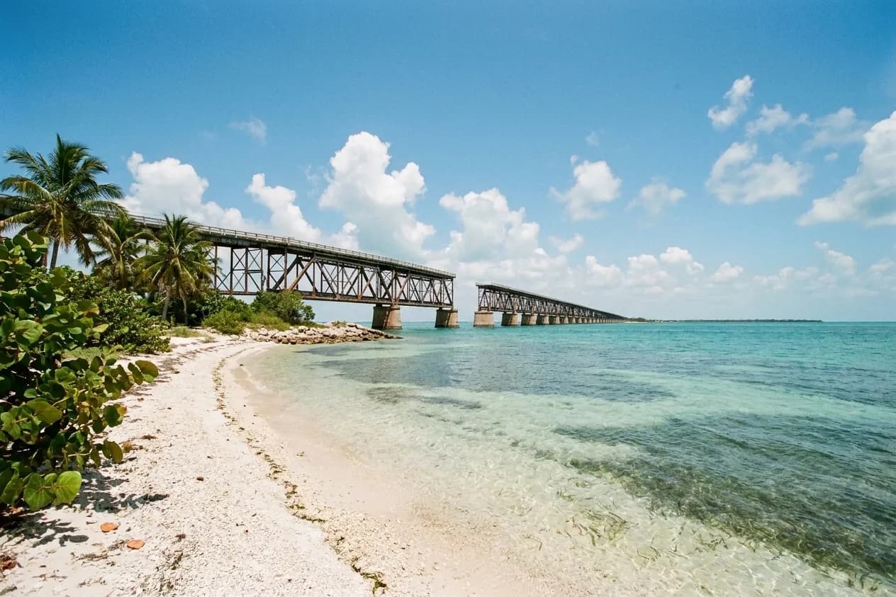 Late morning in the Florida Keys, a pale coarse-sand beach curving away past clear turquoise shallows with the rusted trusses of an old railway bridge crossing the channel beyond