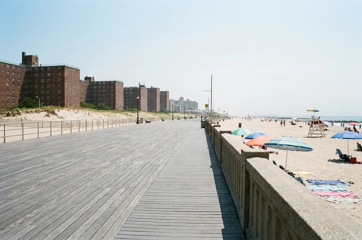 A weathered New York ocean boardwalk on a summer afternoon with umbrellas on the sand and a lifeguard chair beyond