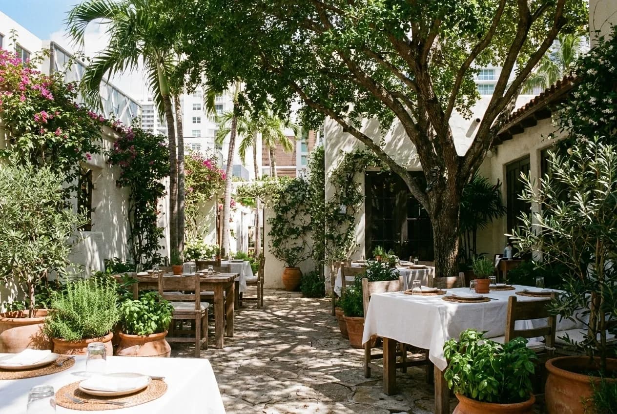 A Mediterranean restaurant courtyard in Miami set for lunch service under a shade tree with dappled sunlight.
