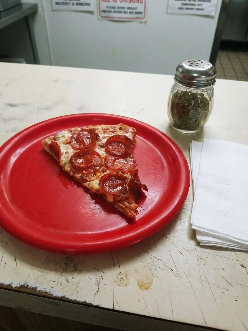 A large plain cheese slice on a paper plate on a formica counter with a shaker of oregano and a stack of napkins
