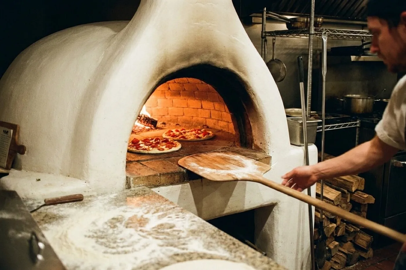 A pizza oven with two pies inside and a peel resting against the opening in a small takeaway kitchen