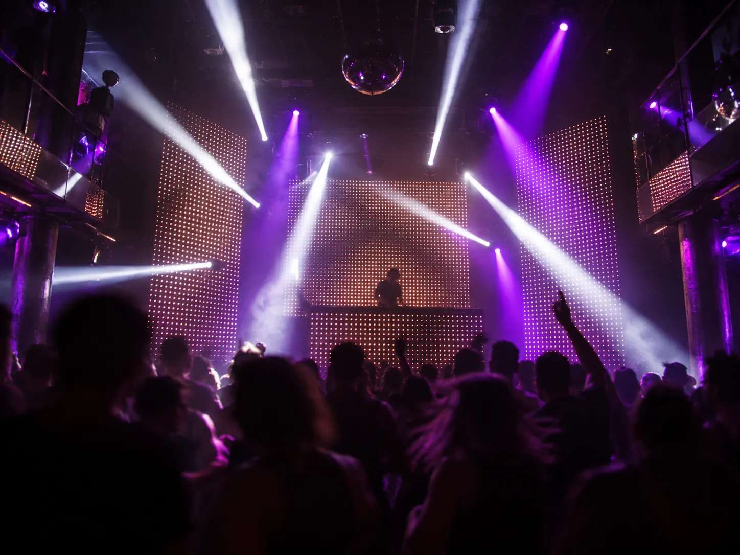 A dark club interior with tall LED screens and beams of light over a dance floor