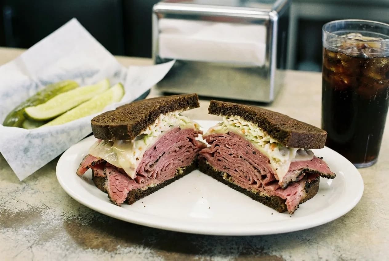A hand-cut pastrami sandwich on dark rye cut in half on a plain white plate at a deli counter