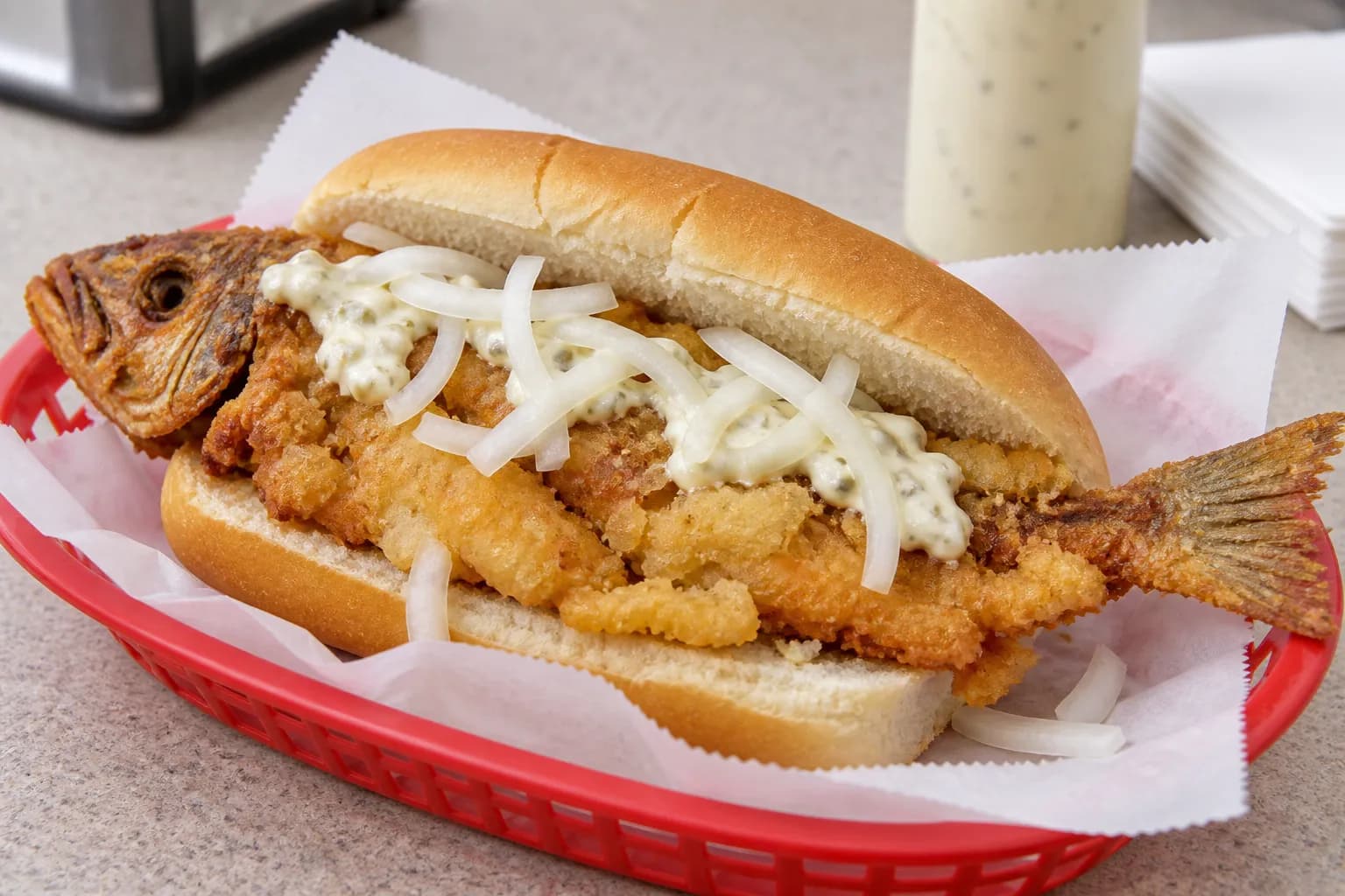 A whole butterflied fried fish folded into a soft roll with raw onion and a squeeze of sauce, the tail sticking out past the bread, on a paper-lined tray