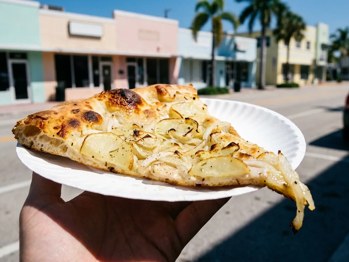 A folded slice of pizza on a paper plate held up in bright Miami daylight with pastel buildings blurred behind