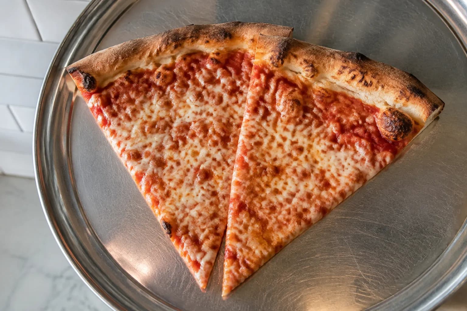 Fresh New York pizza slices on a metal tray at a counter, viewed from above