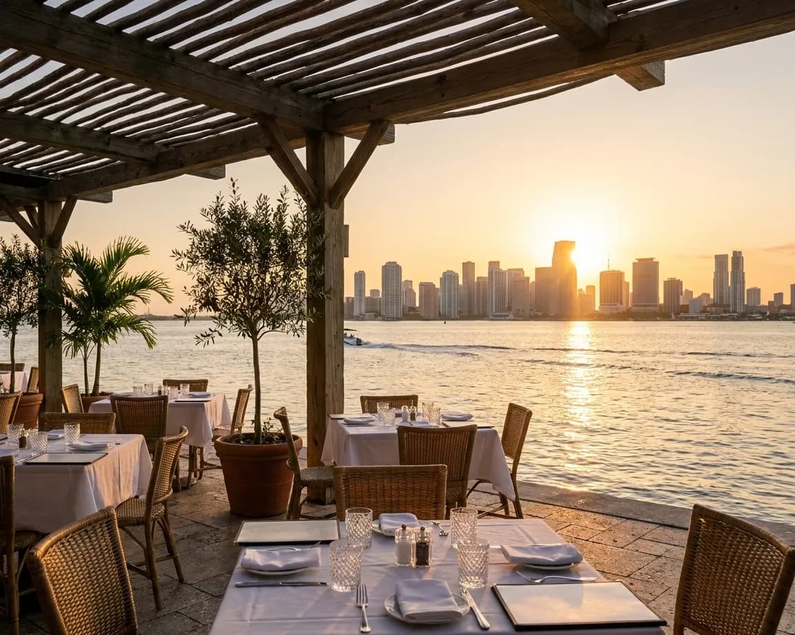 A bayfront restaurant terrace in Miami at golden hour, linen-set tables under a pergola with the skyline on the horizon