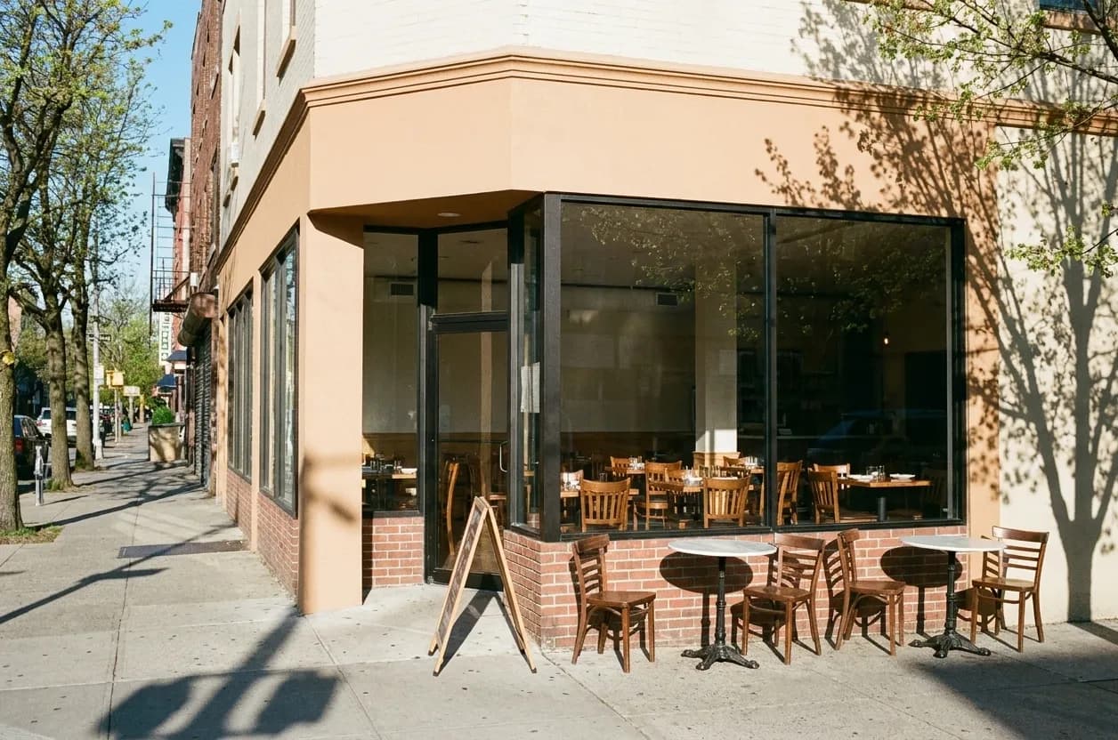 A newly painted Brooklyn restaurant storefront on a bright weekday morning, plate glass showing the room inside and two unset outdoor tables on the pavement