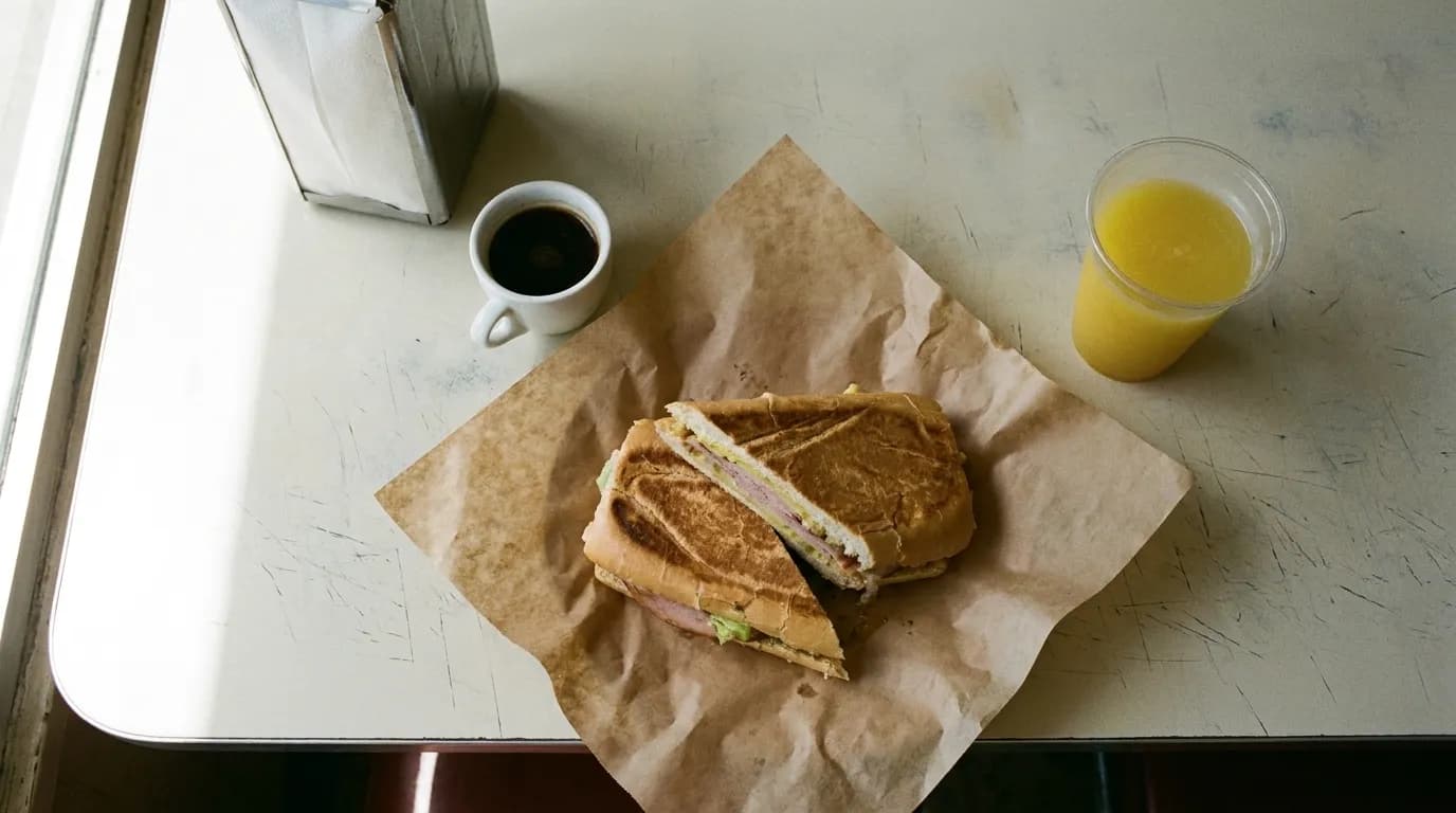 An overhead view of a Cuban lunch counter with a pressed sandwich on butcher paper, a small cup of dark coffee and a napkin dispenser