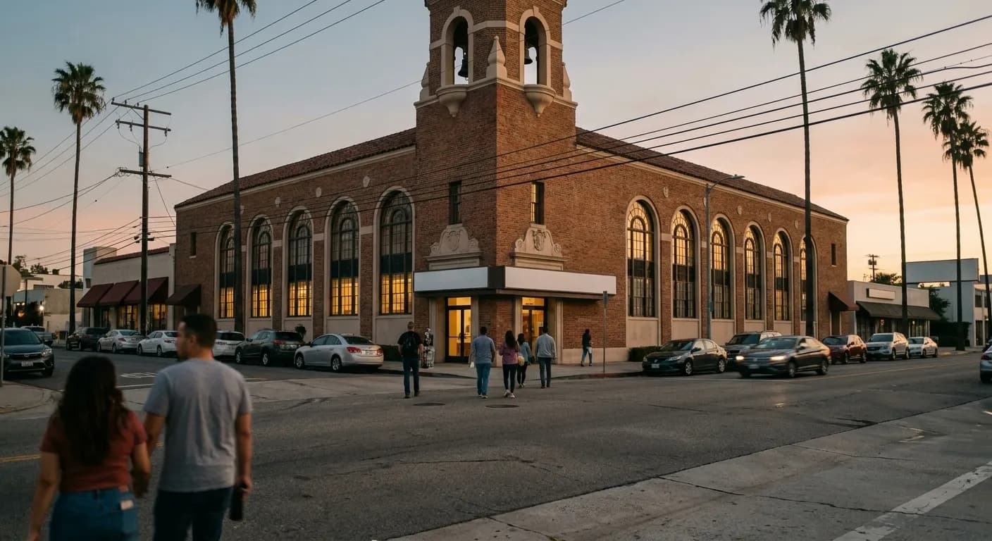 A low-rise Los Angeles boulevard at dusk, a 1929 brick building with tall arched windows glowing warm as a few people walk toward the entrance