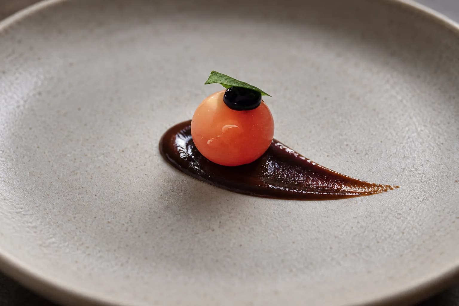 A single tiny composed bite, a glossy translucent sphere on a smear of dark sauce, alone at the centre of an oversized matte plate