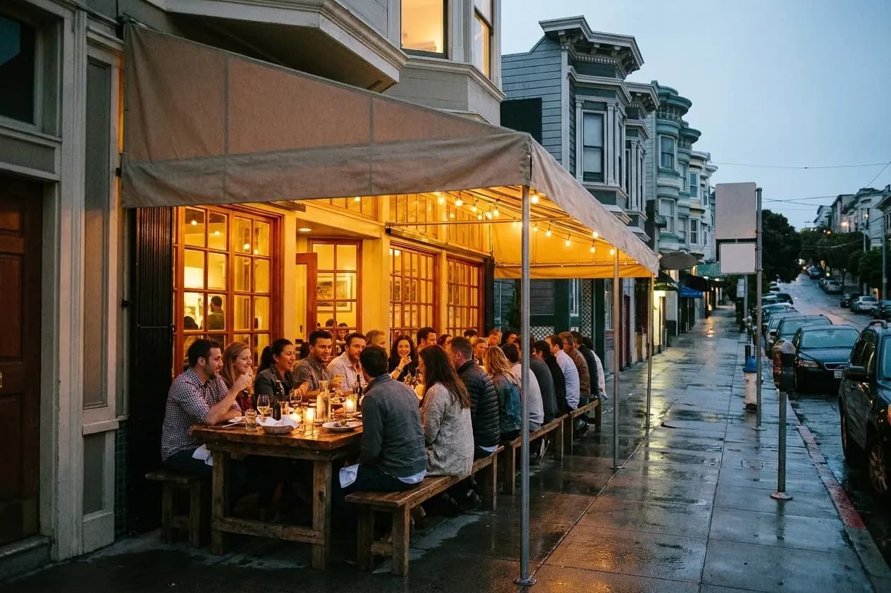 A long communal table of a large group dining under an awning outside a warmly lit San Francisco restaurant at dusk