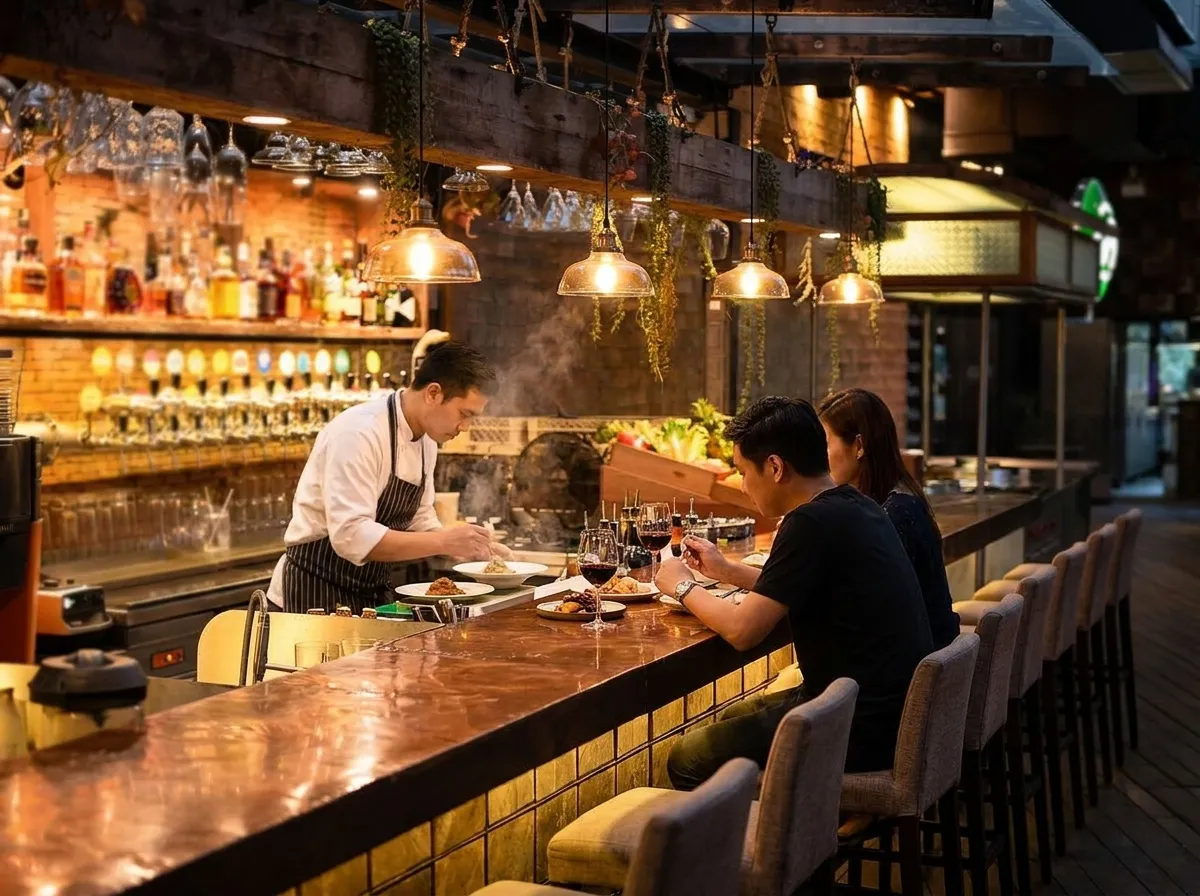 A warm restaurant bar counter at dinner service with a row of stools, solo diners eating small plates and a cook plating behind the pass