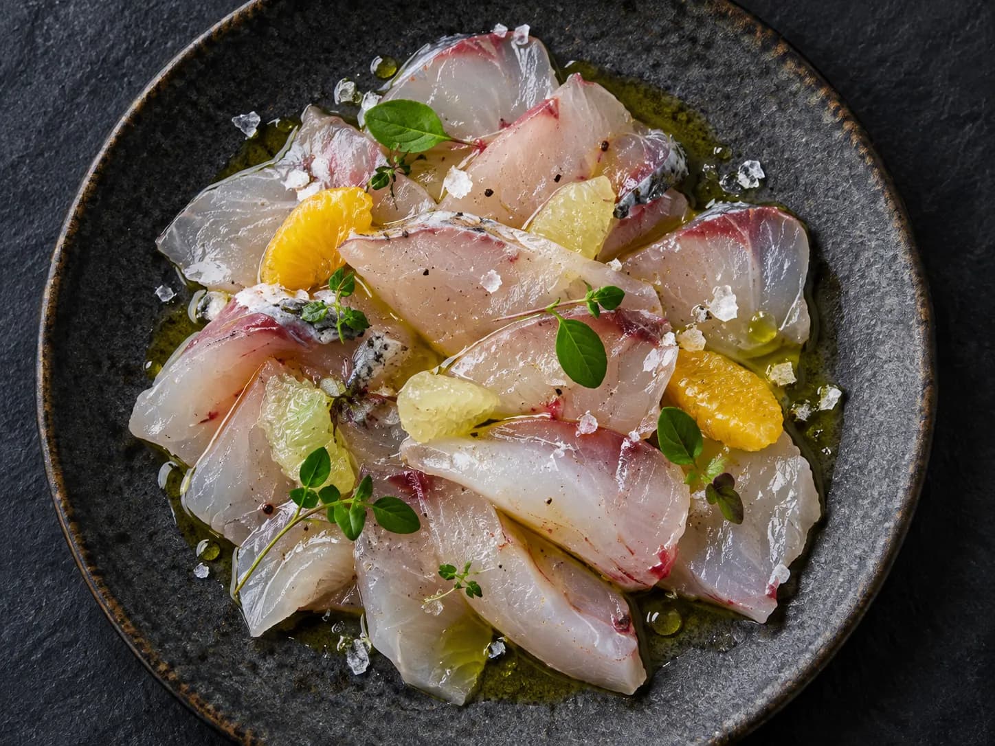 Thin slices of raw fish arranged on a chilled plate with citrus, oil and a scattering of herbs