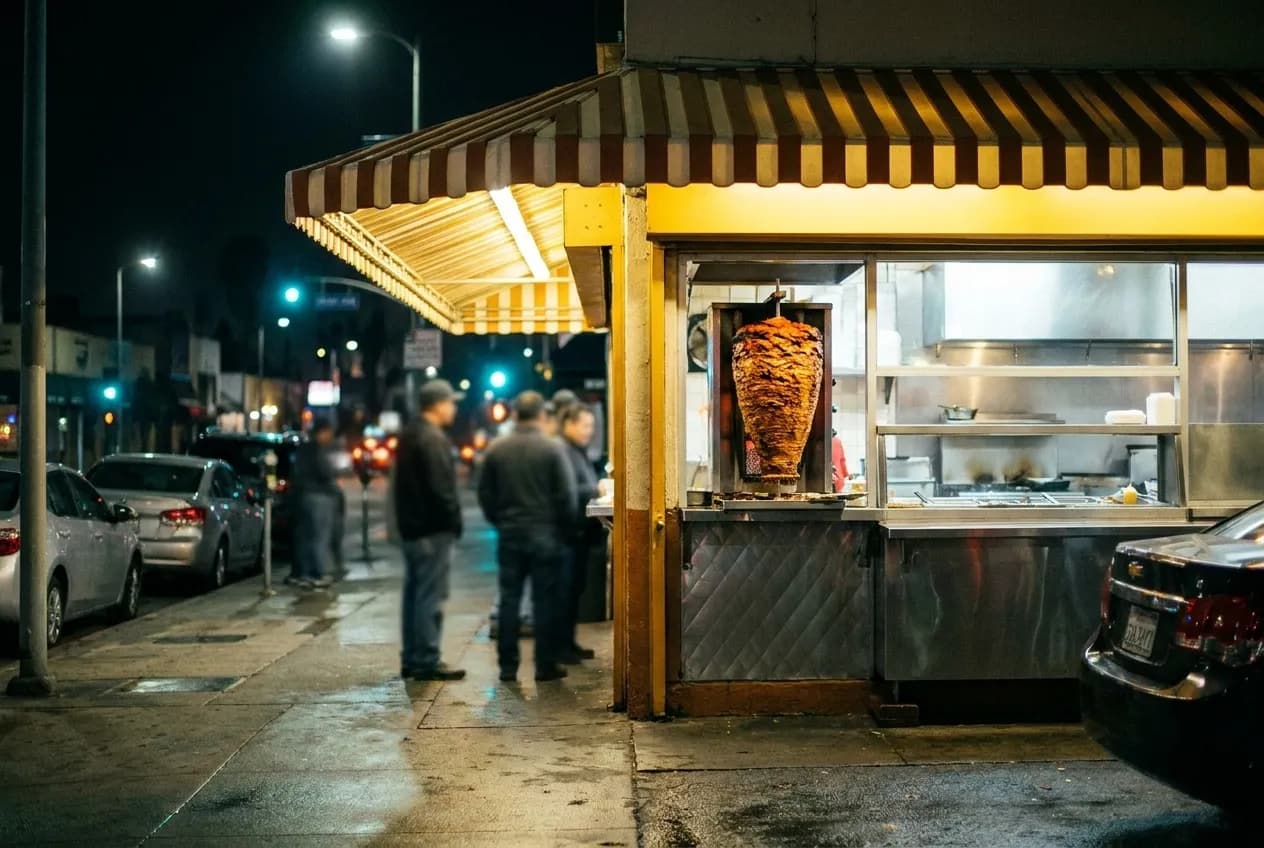 Night view of a Los Angeles taco stand with vertical trompo spit and customers waiting under incandescent lights