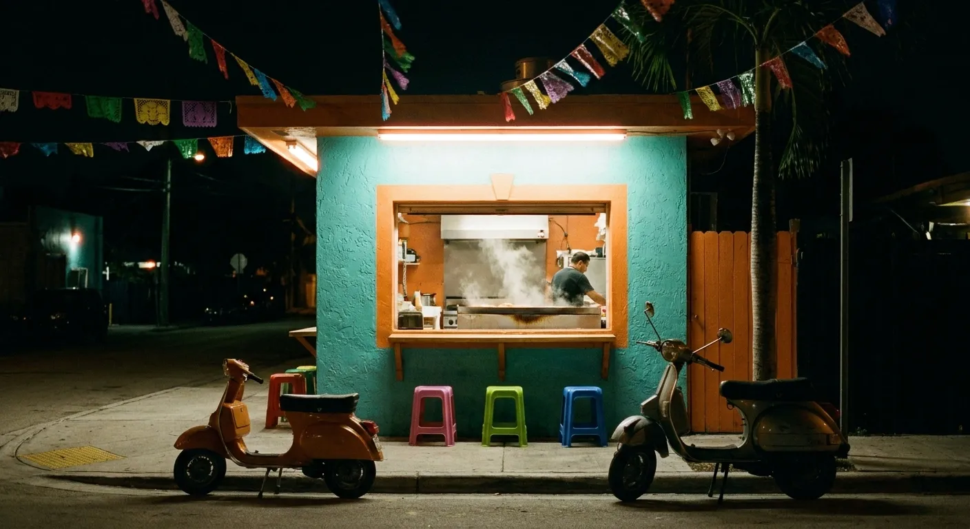 A small brightly painted taqueria on a Miami side street at night, turquoise and orange walls around a lit service window with steam rising from the plancha, three stools at a ledge outside