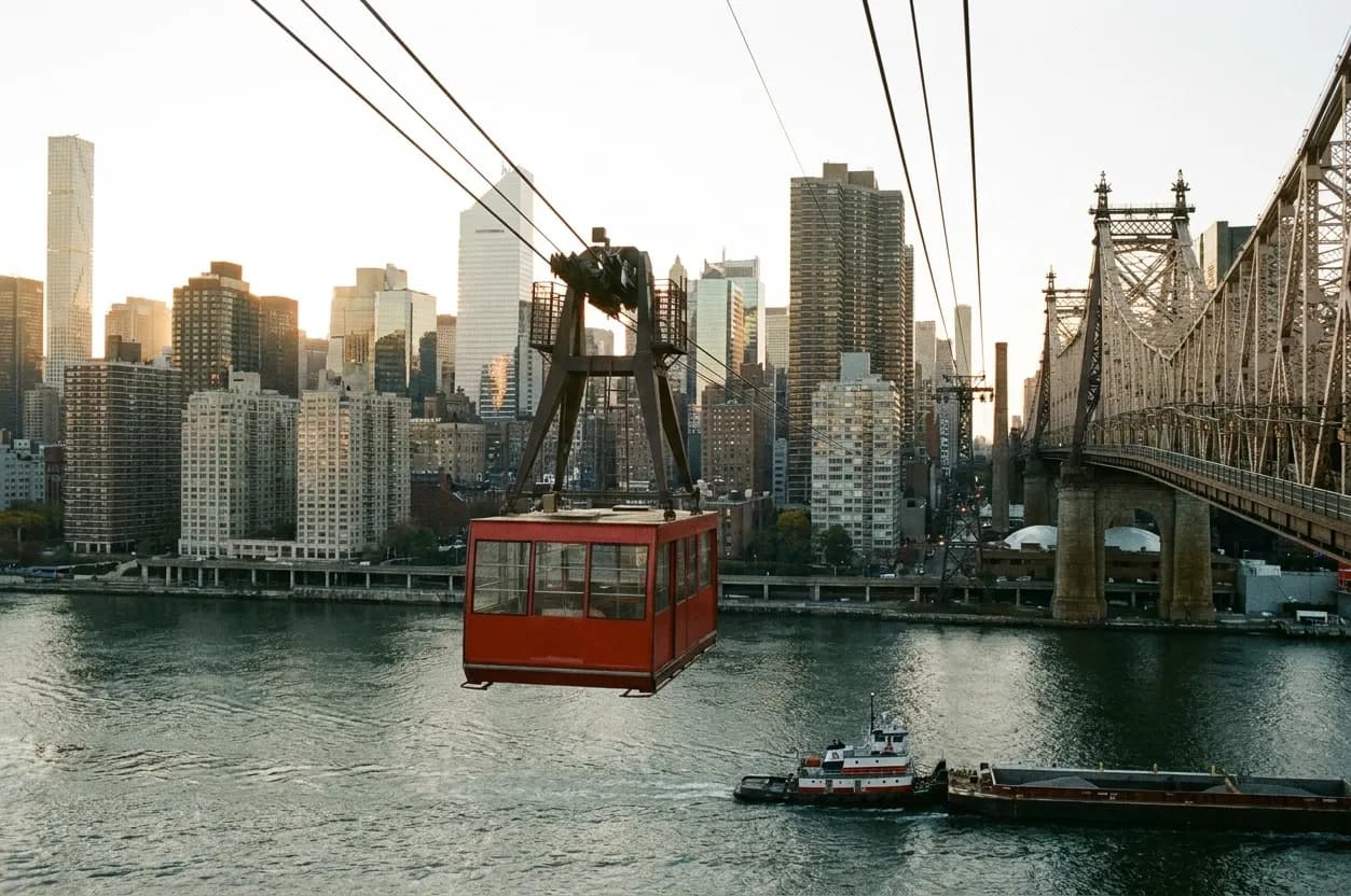 A red aerial tram cabin crossing high above the East River beside a steel bridge with Midtown behind