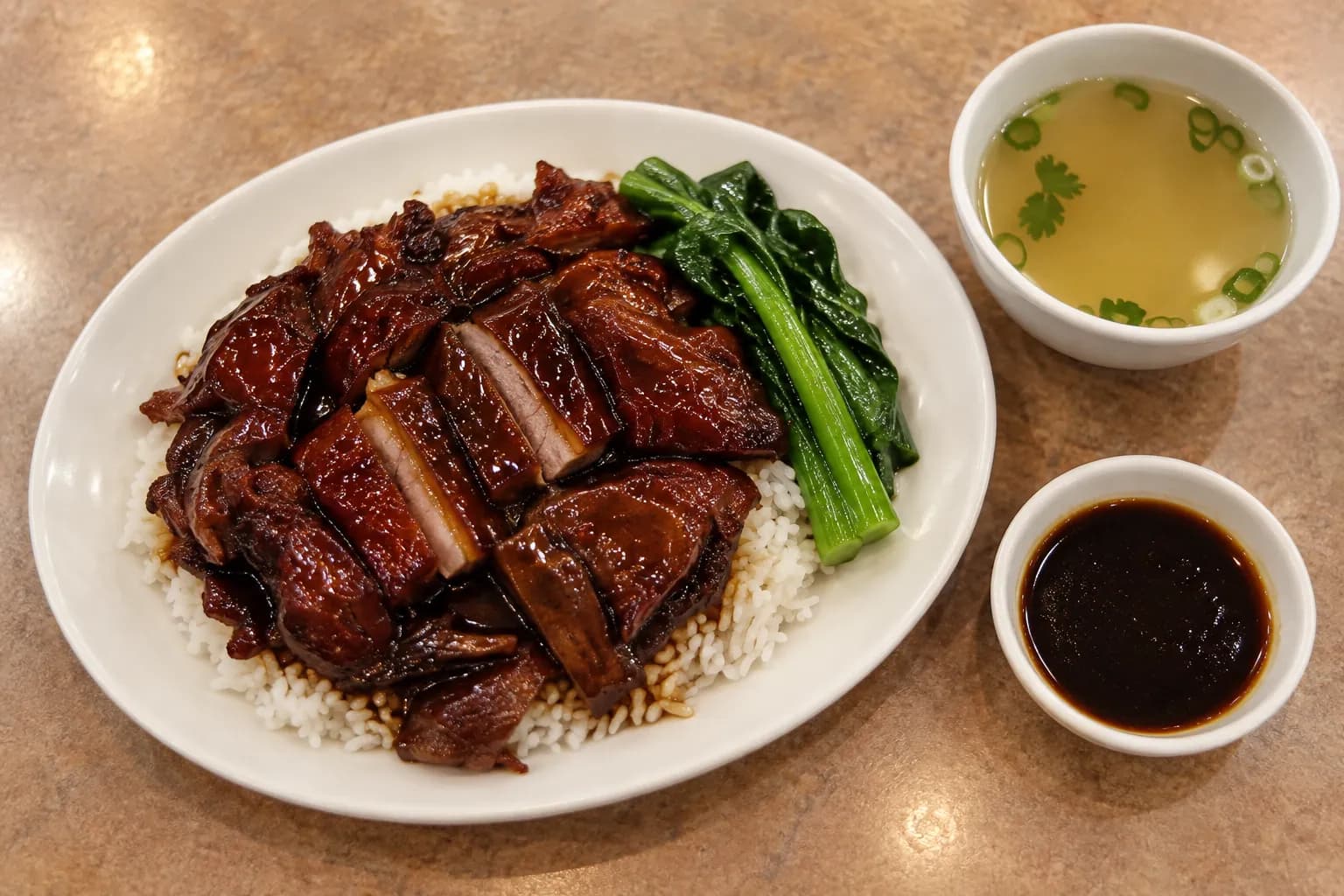 An oval plate of sliced lacquered roast meat over white rice with blanched greens and a dish of dark sauce, a bowl of clear broth beside