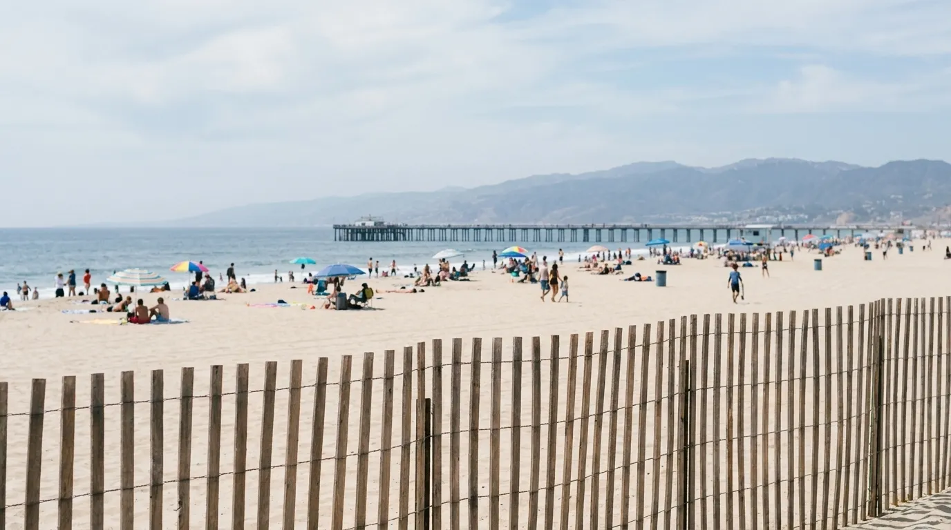 A wide Los Angeles beach in the afternoon with a pier in the distance, towels and umbrellas spread across pale sand and the ocean flat behind