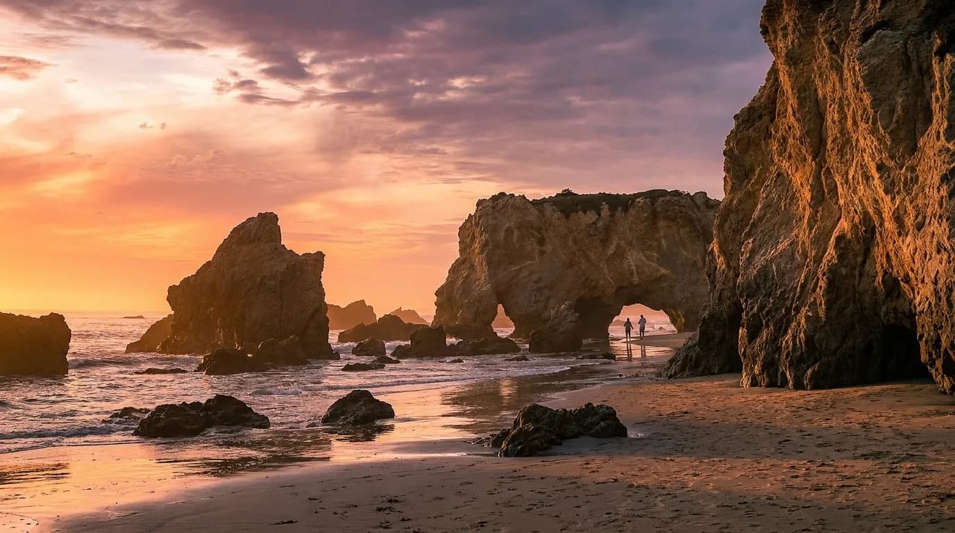 A narrow Malibu cove at low tide with sea stacks and eroded rock arches, a few figures far along the sand and low golden light coming in under the cliff